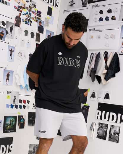 A man wearing a black RUDIS t-shirt and white shorts, standing in front of a white wall covered with fashion design sketches and apparel displays.