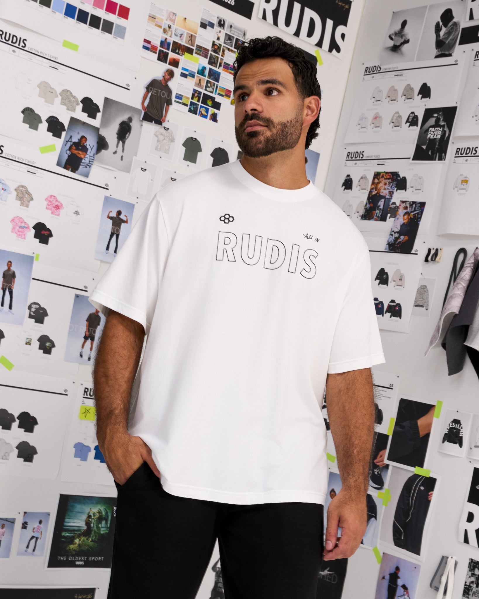 A man wearing a white oversized t-shirt with 'RUDIS' printed across the chest, standing in front of a white wall covered in fashion sketches and design boards.