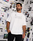 A man wearing a white oversized t-shirt with 'RUDIS' printed across the chest, standing in front of a white wall covered in fashion sketches and design boards.