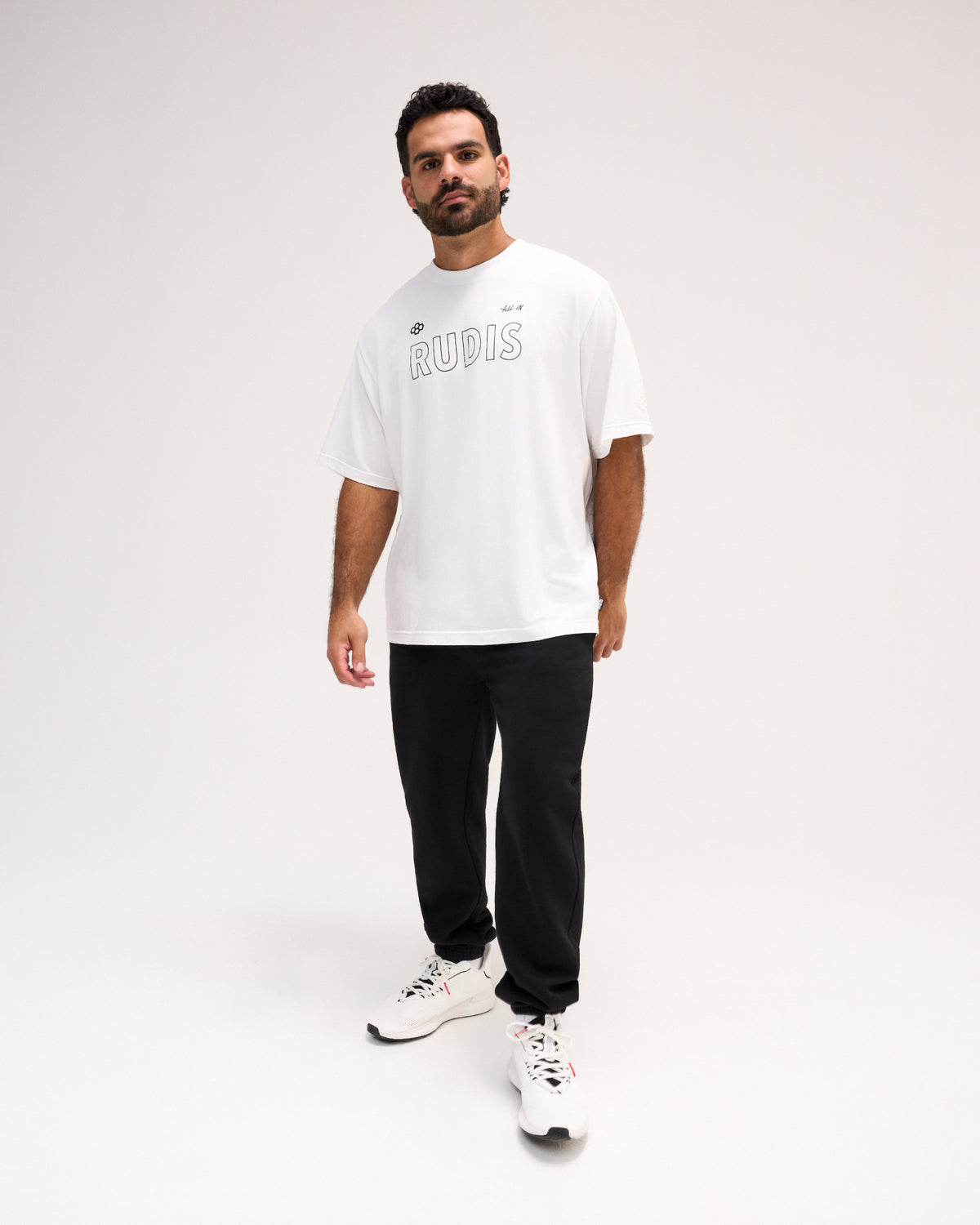 A man modeling a white oversized t-shirt with 'RUDIS' printed in gray outline text on the chest, paired with black pants and white sneakers, against a plain light gray studio background.