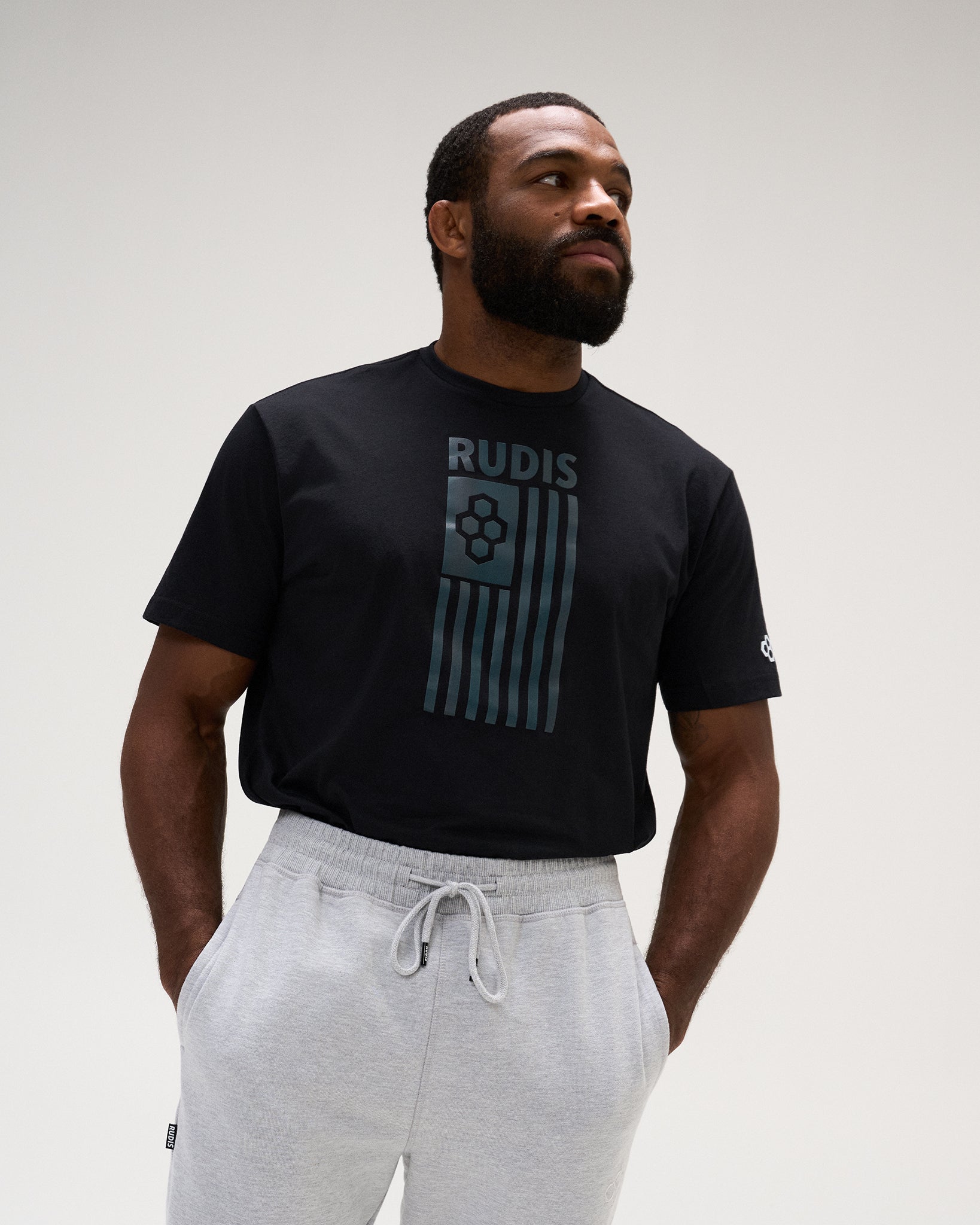 A man wearing a black short-sleeve t-shirt with a dark teal graphic on the front that reads 'RUDIS' above vertical stripes, paired with light gray sweatpants, against a plain light gray studio background.