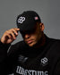 Person wearing a black cap and sweatshirt with logos on a gray background