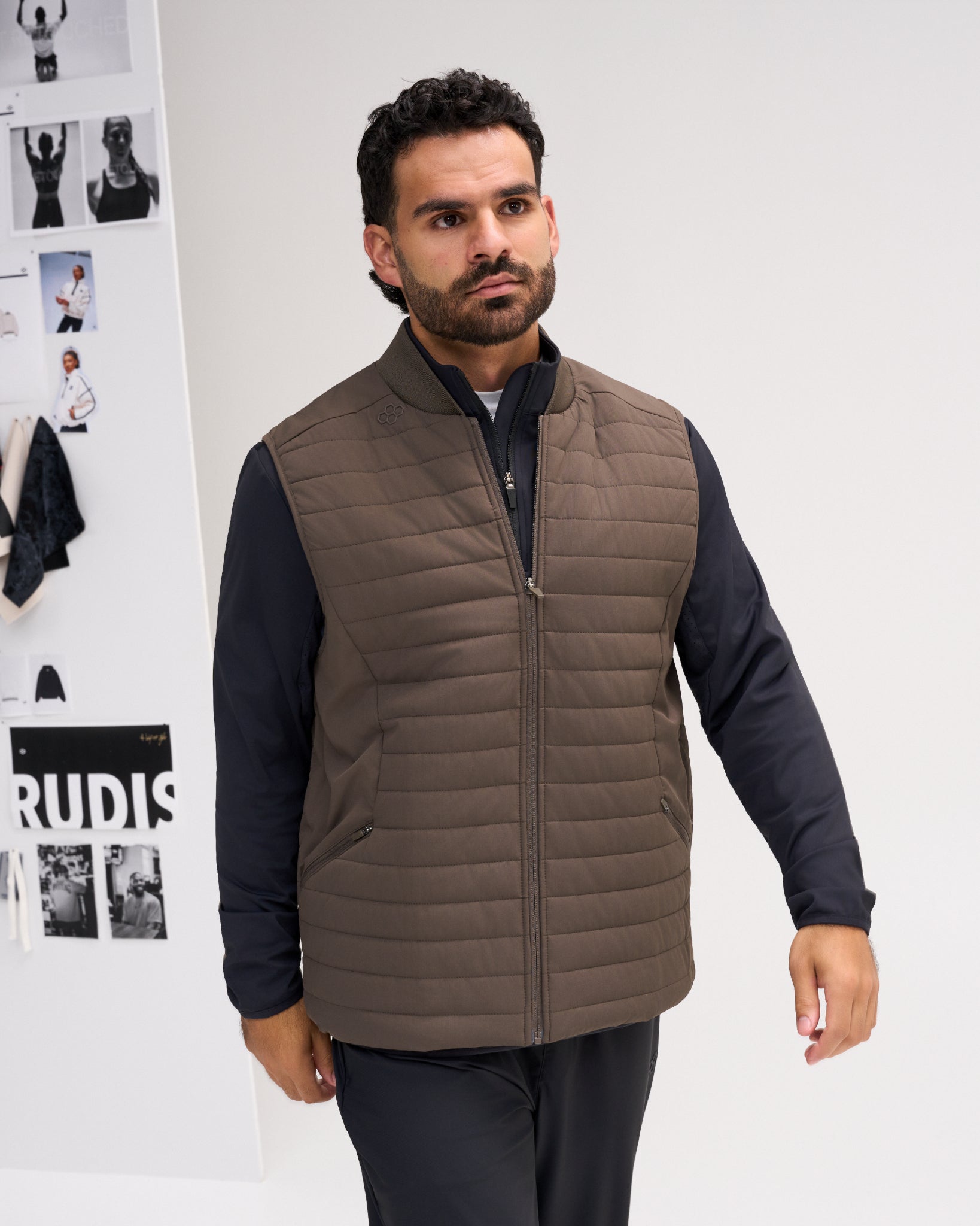A man wearing a brown quilted zip-up vest over a black long-sleeve shirt and black trousers, standing against a white wall with fashion magazine covers in the background.