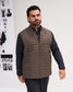 A man wearing a brown quilted zip-up vest over a black long-sleeve shirt and black trousers, standing against a white wall with fashion magazine covers in the background.