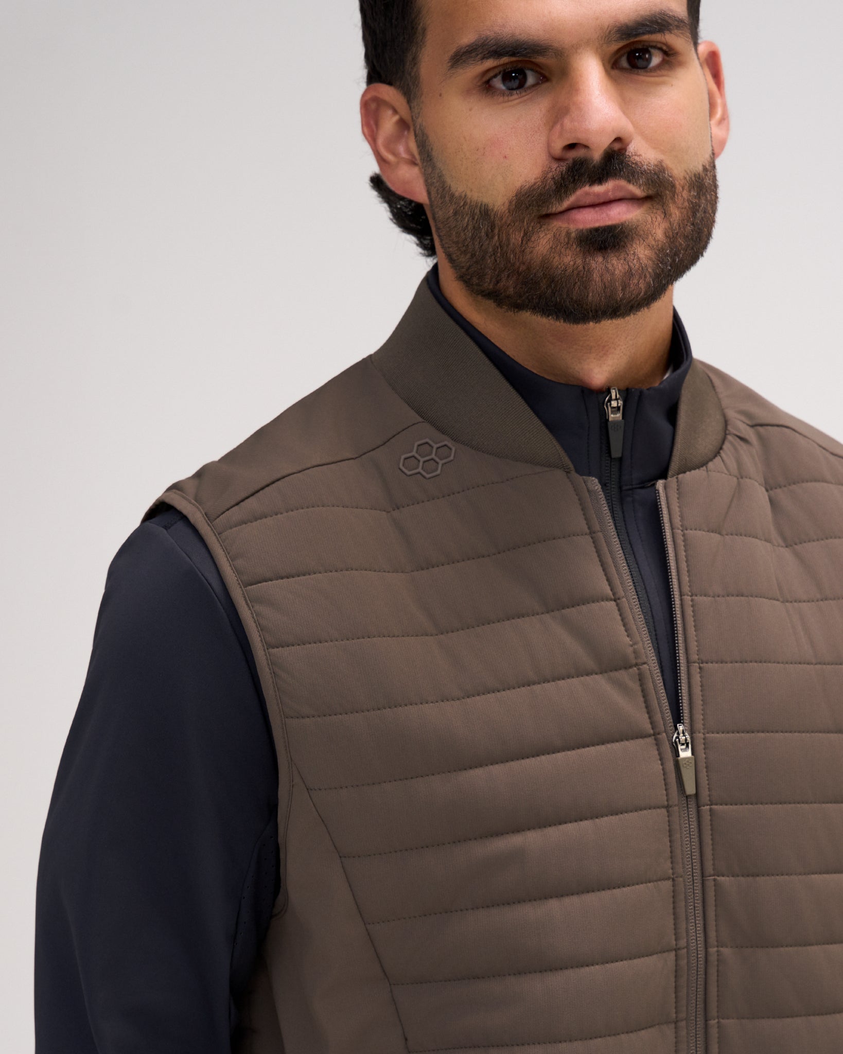 A close-up of a man wearing a taupe brown quilted zip-up vest over a dark navy shirt, against a light gray studio background.