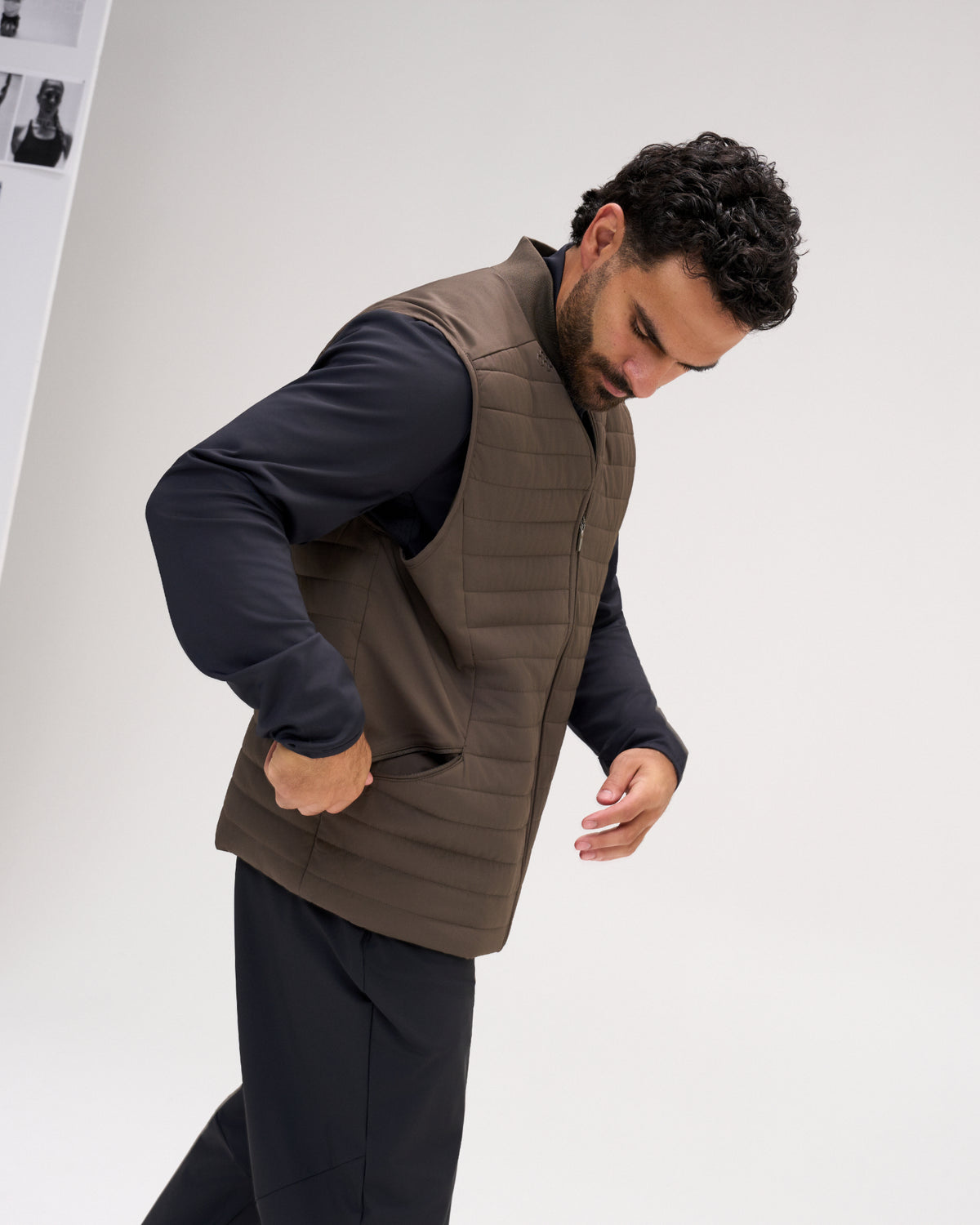 A man wearing a brown quilted puffer vest over a black long-sleeve shirt and black pants, captured in a studio setting against a light gray background.