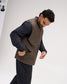 A man wearing a brown quilted puffer vest over a black long-sleeve shirt and black pants, captured in a studio setting against a light gray background.