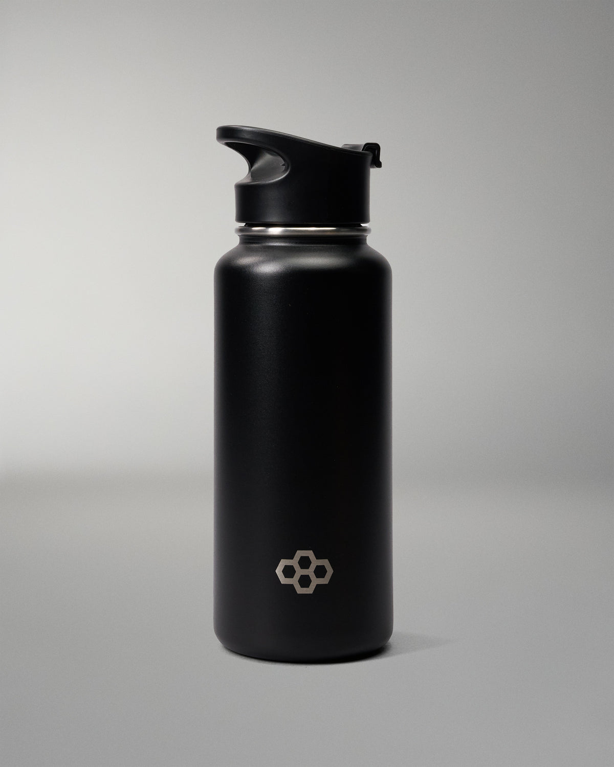 A matte black insulated water bottle with a matching black flip-top lid, featuring a small silver geometric logo near the bottom, set against a neutral gray studio background.
