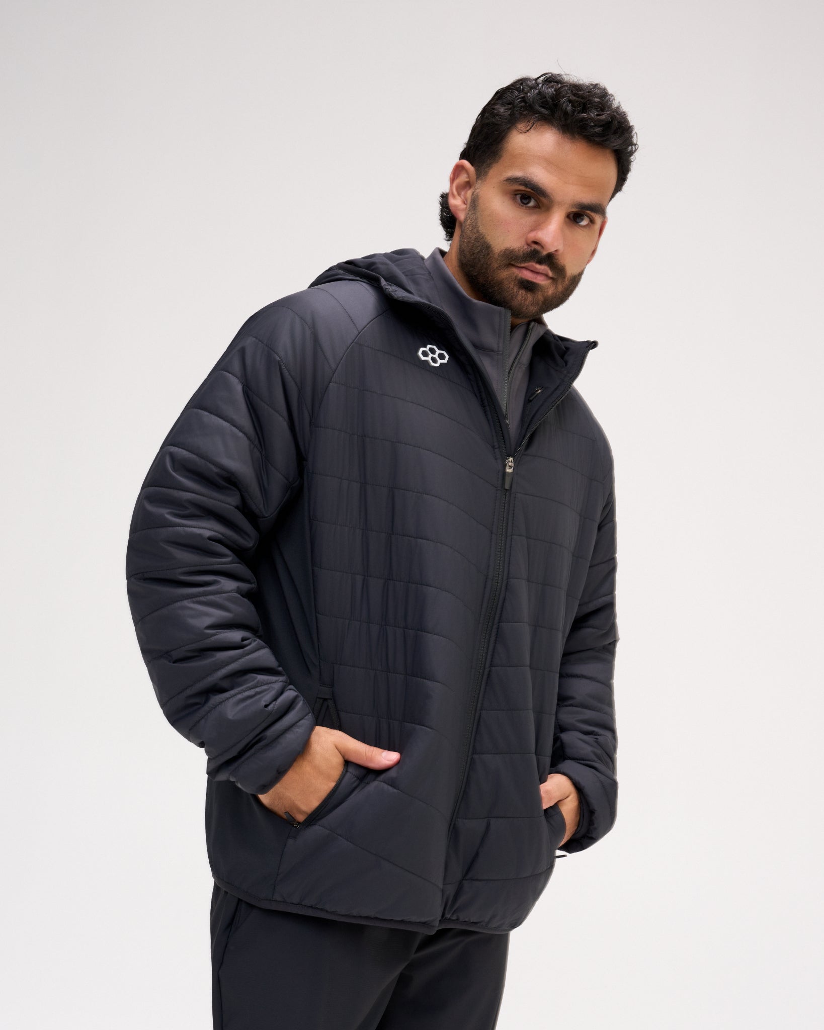A man wearing a Black quilted puffer jacket with a white geometric logo on the chest, standing against a light gray studio background.