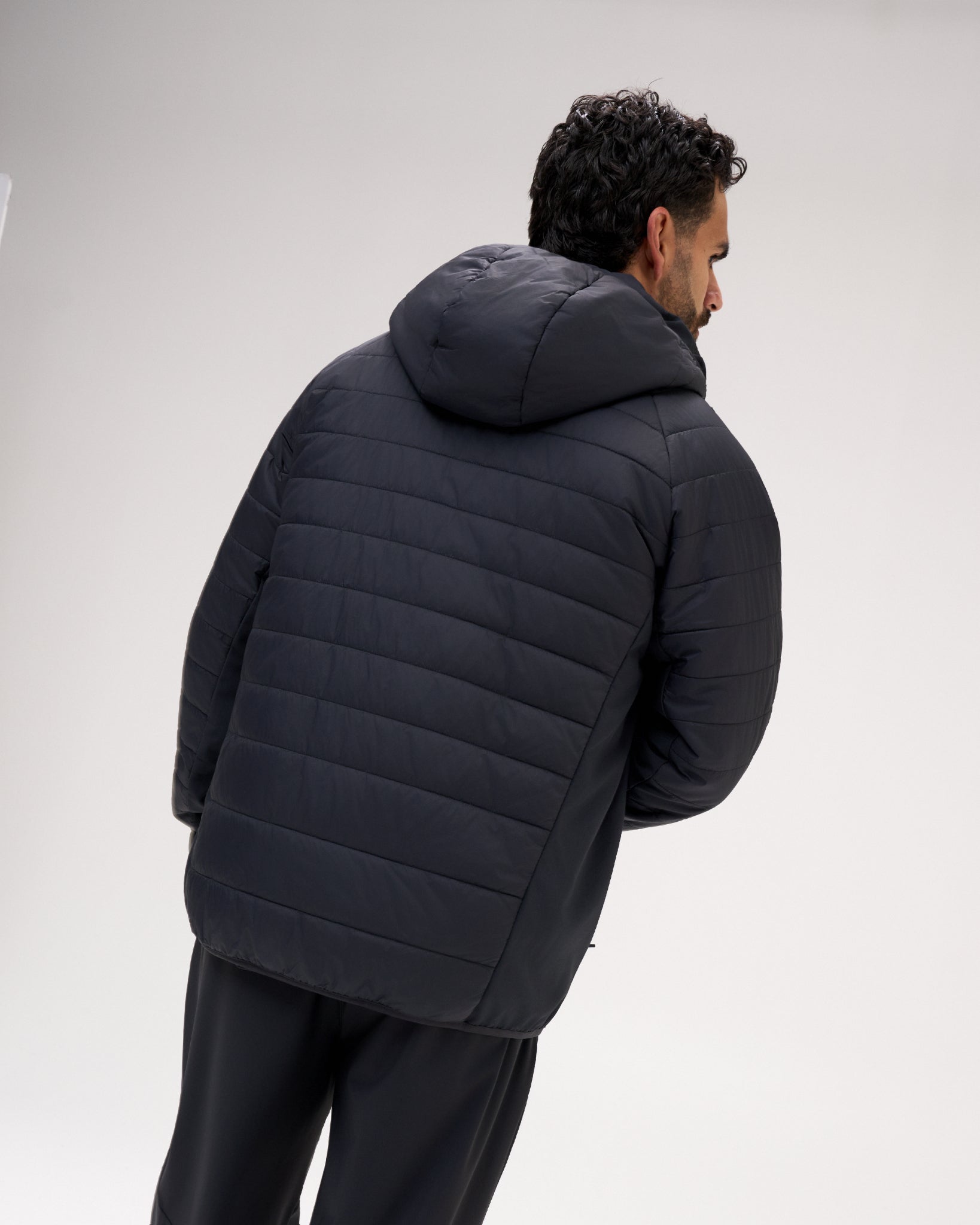 A man wearing a black quilted puffer jacket with a hood, shown from a rear three-quarter angle against a light gray studio background.