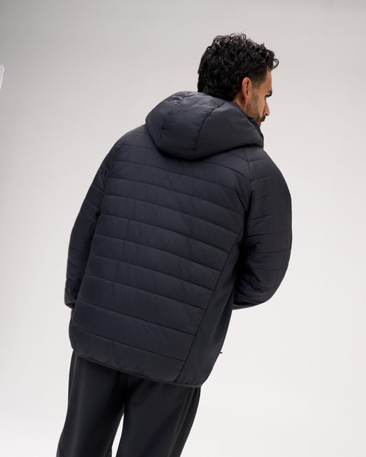 A man wearing a black quilted puffer jacket with a hood, shown from a rear three-quarter angle against a light gray studio background.