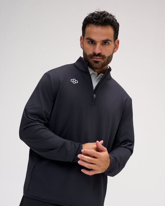 A man wearing a dark navy long-sleeve quarter-zip pullover with a small white logo on the left chest, layered over a light gray collared shirt, against a plain light gray studio background.