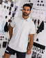 A man wearing a white short-sleeve polo shirt and black shorts, standing in front of a wall covered with fashion sketches and apparel photos. The brand name RUDIS is visible on the wall and on the shorts.