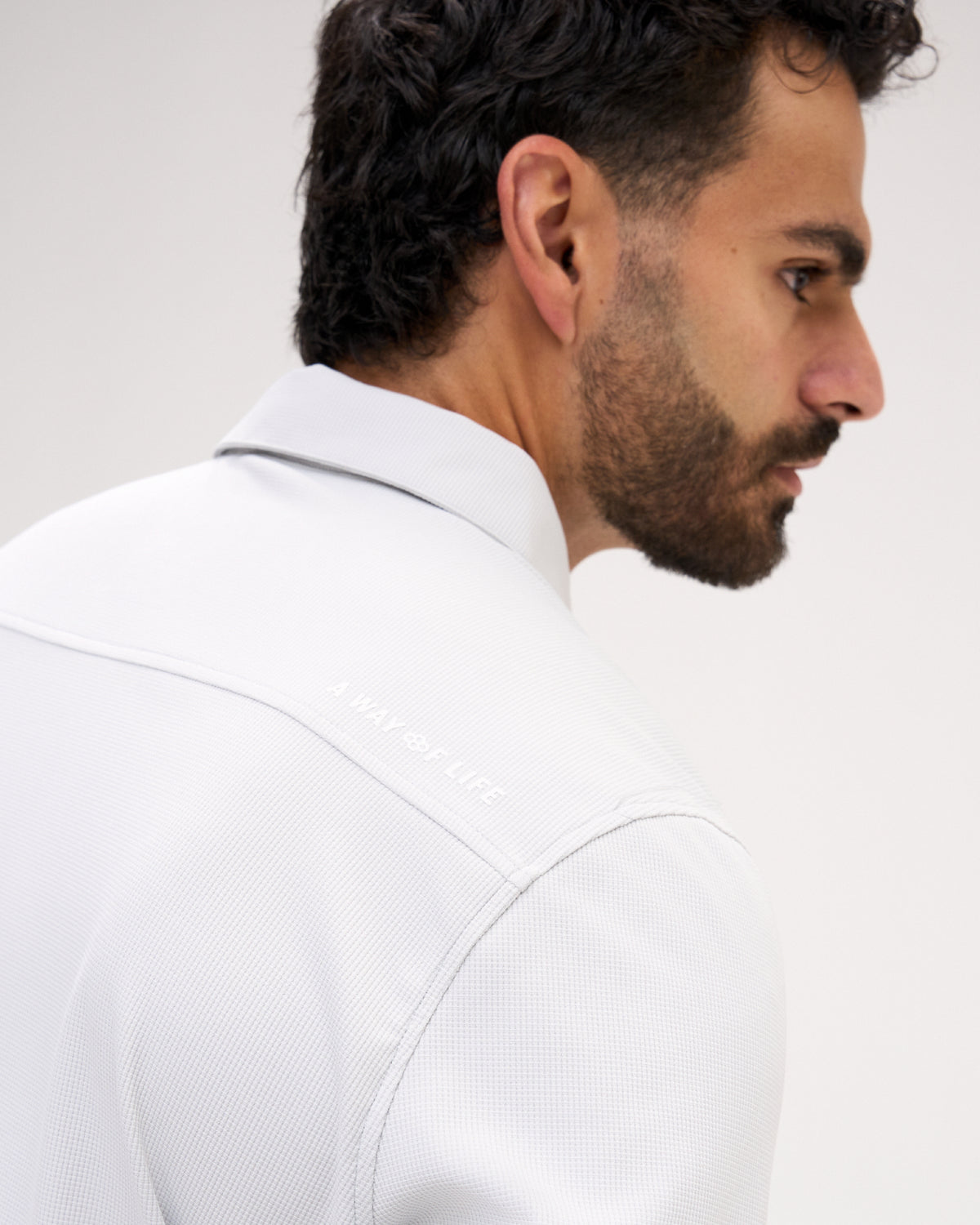 A close-up side profile of a man wearing a white textured collared shirt, showing the shoulder and upper back with subtle tonal embroidery.