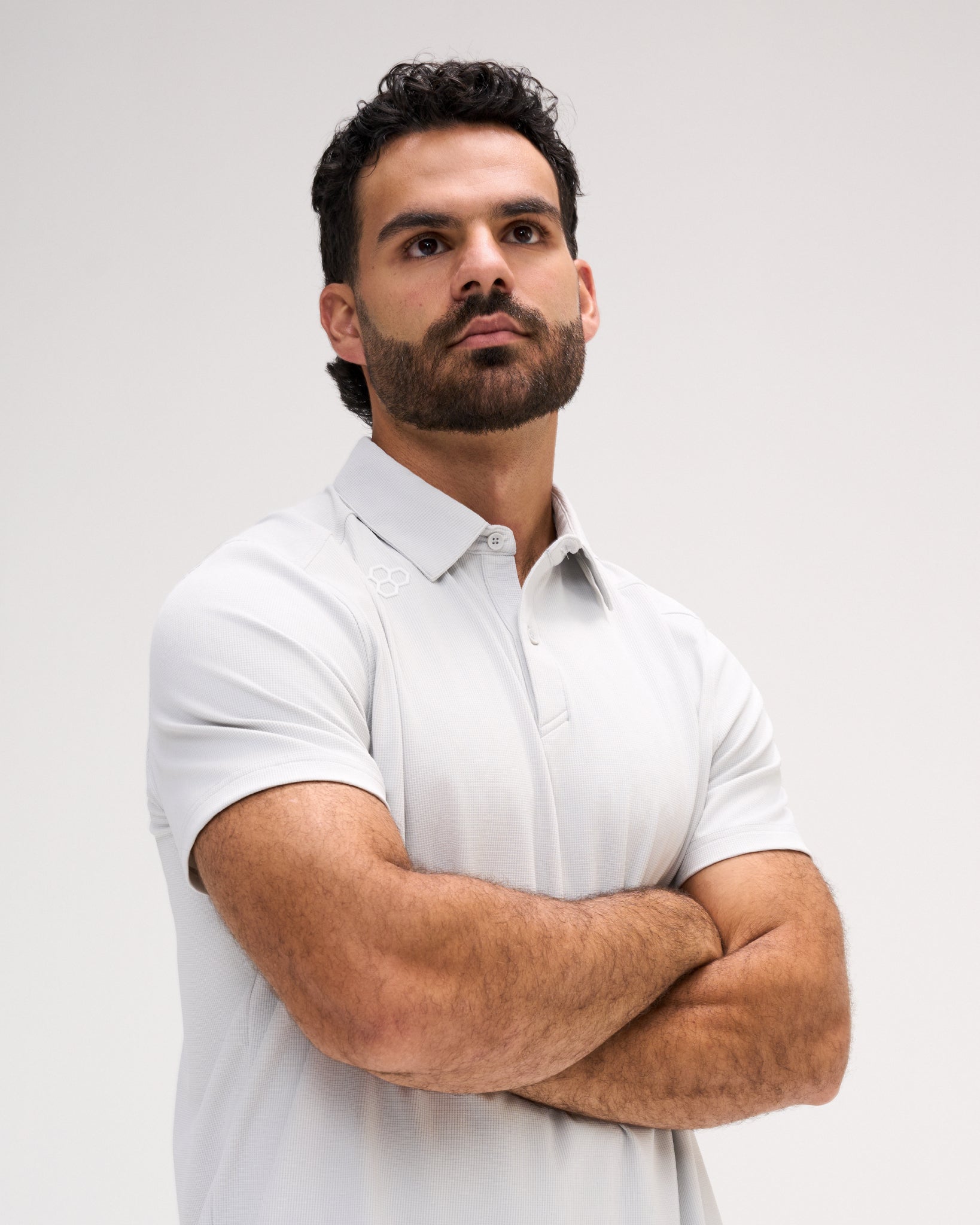 Male model wearing the RUDIS Go-To Polo in Lunar Gray, featuring a subtle textured fabric, short sleeves, a classic button placket, structured collar, and tonal RUDIS hex logo near the right shoulder, posed with arms crossed.