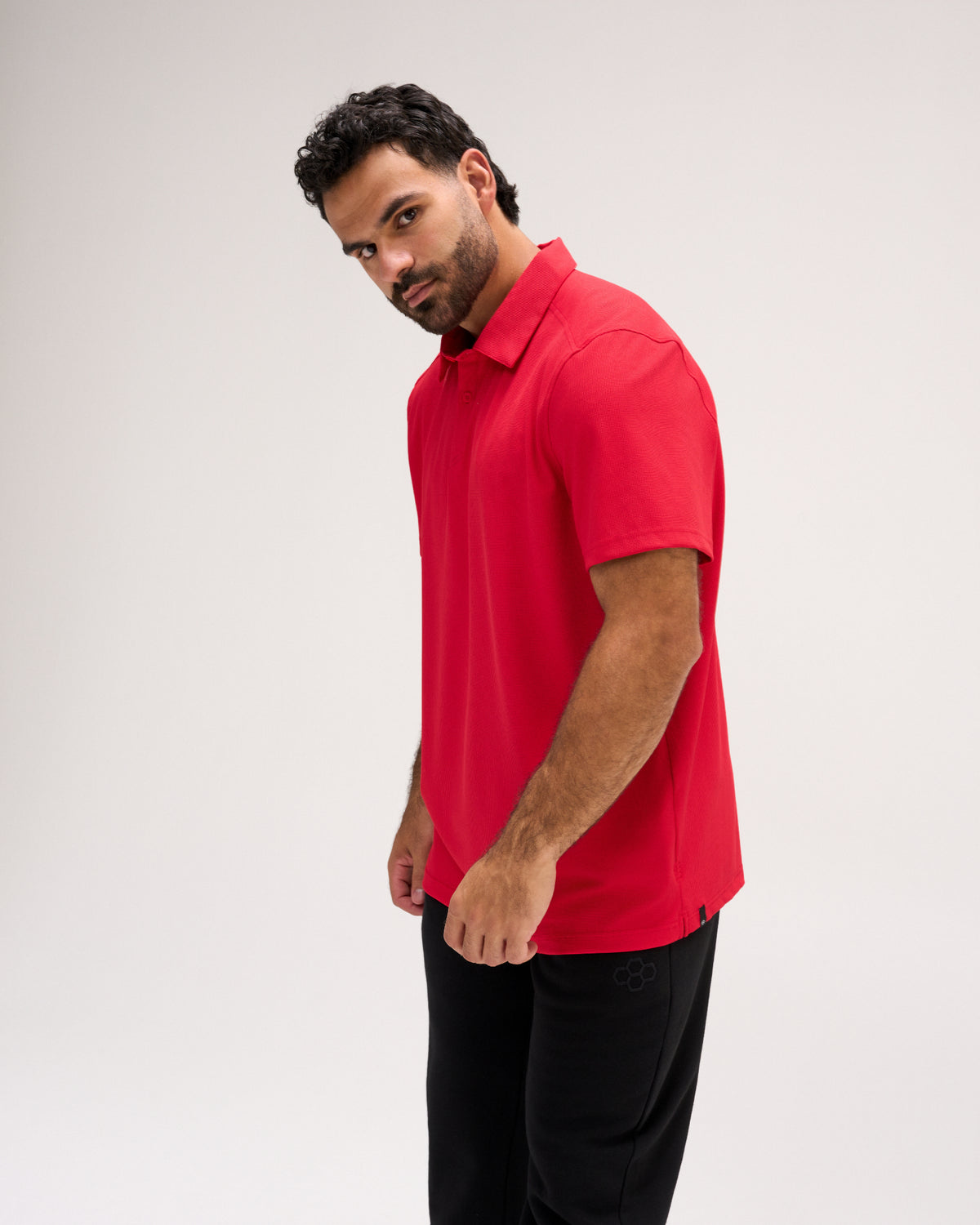 Male model wearing the RUDIS Go-To Polo in Red with black athletic pants, showcasing the short-sleeve design, structured collar, and relaxed athletic fit from a three-quarter side view.