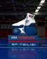 White and blue wrestling shoes on a blue platform with 'USA Wrestling' and 'SF-Tbilisi' text, in an indoor sports facility.