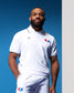 An adult male model wearing a white USA Wrestling athletic polo and shorts, standing on a red pedestal with a blue and white background.