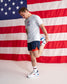 A man in athletic wear stretches his leg against a large American flag backdrop. He is wearing a light gray USA-themed t-shirt, navy athletic shorts with red side panels, white socks, and white running shoes with blue and red accents.