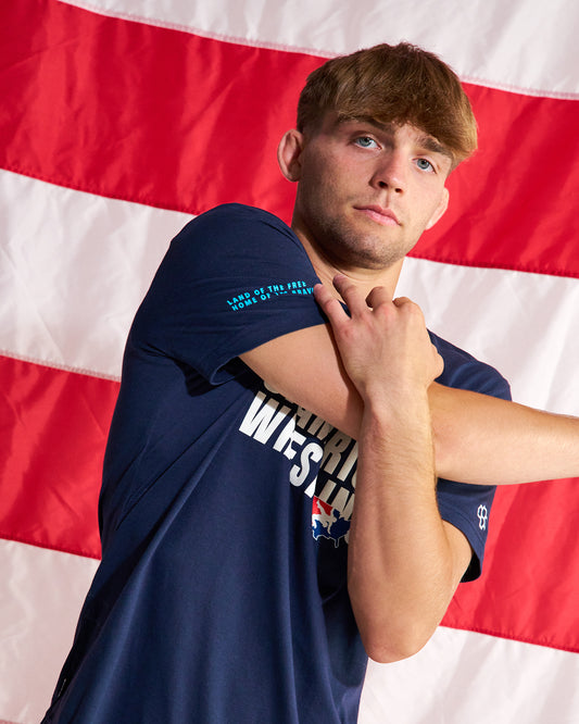 A young man wearing a navy blue athletic t-shirt with 'UNITED STATES OF AMERICA' printed on the front, posing in front of a red and white American flag backdrop.