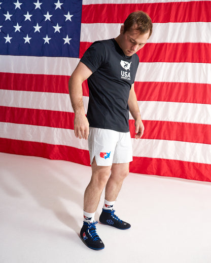 A man wearing a black USA boxing team t-shirt, white boxing shorts, and blue boxing shoes stands in front of a large American flag backdrop.