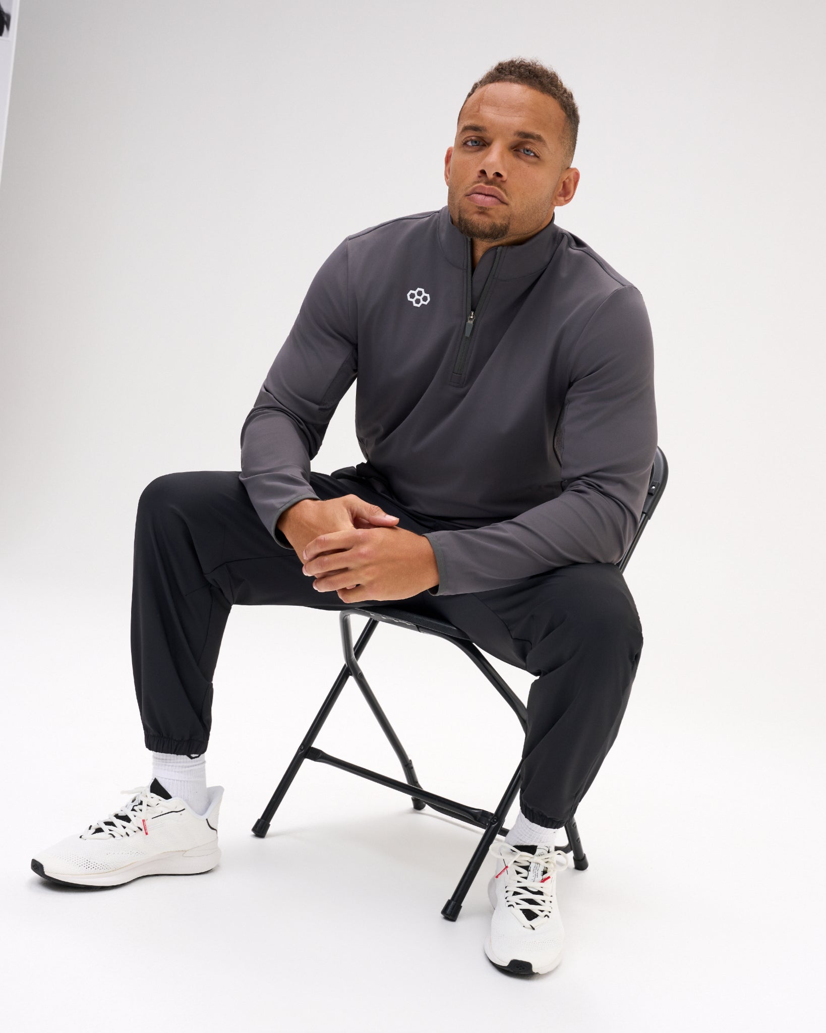 Seated male model wearing the RUDIS Victory 1/4 Zip in Carbon with black athletic pants, showcasing the quarter-zip neckline, white RUDIS chest logo, and relaxed athletic styling.