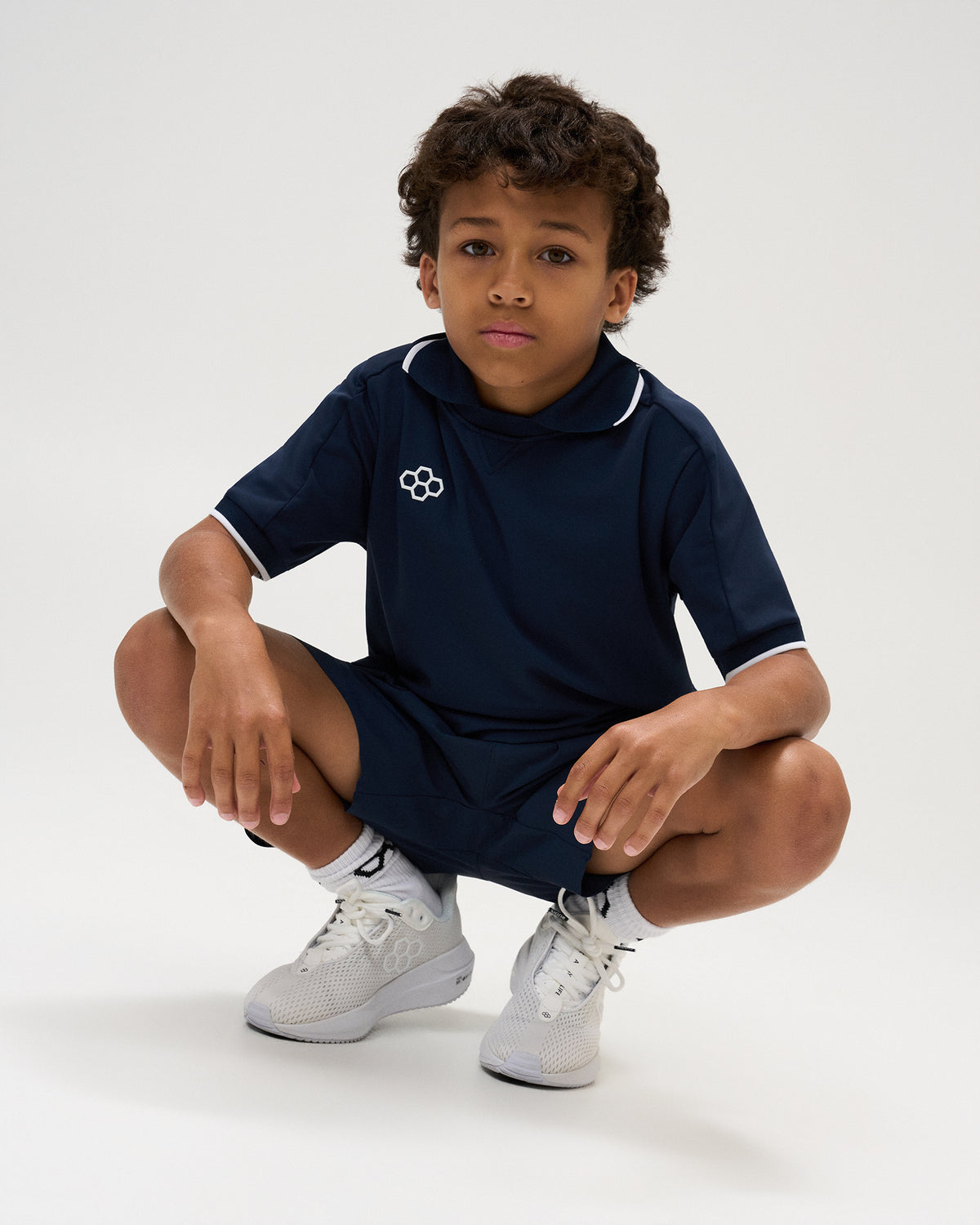 Youth model crouching in the RUDIS Chronicle Elite Youth Athletic Polo in Navy, paired with matching navy athletic shorts and white sneakers, showcasing the white RUDIS chest logo and contrast-trimmed collar.