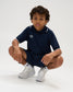 Youth model crouching in the RUDIS Chronicle Elite Youth Athletic Polo in Navy, paired with matching navy athletic shorts and white sneakers, showcasing the white RUDIS chest logo and contrast-trimmed collar.