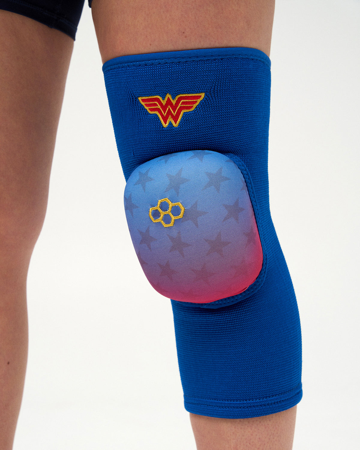 Close-up of RUDIS Wonder Woman Cushion Wrestling Knee Pad with blue knit sleeve, gradient star-patterned padding, gold RUDIS logo and Wonder Woman emblem.