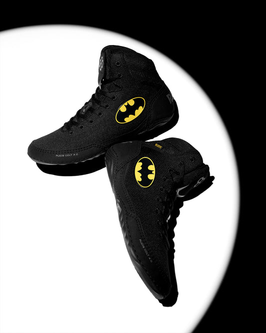 Two black high-top wrestling shoes with yellow Batman logos, floating against a white circular spotlight on a black background.