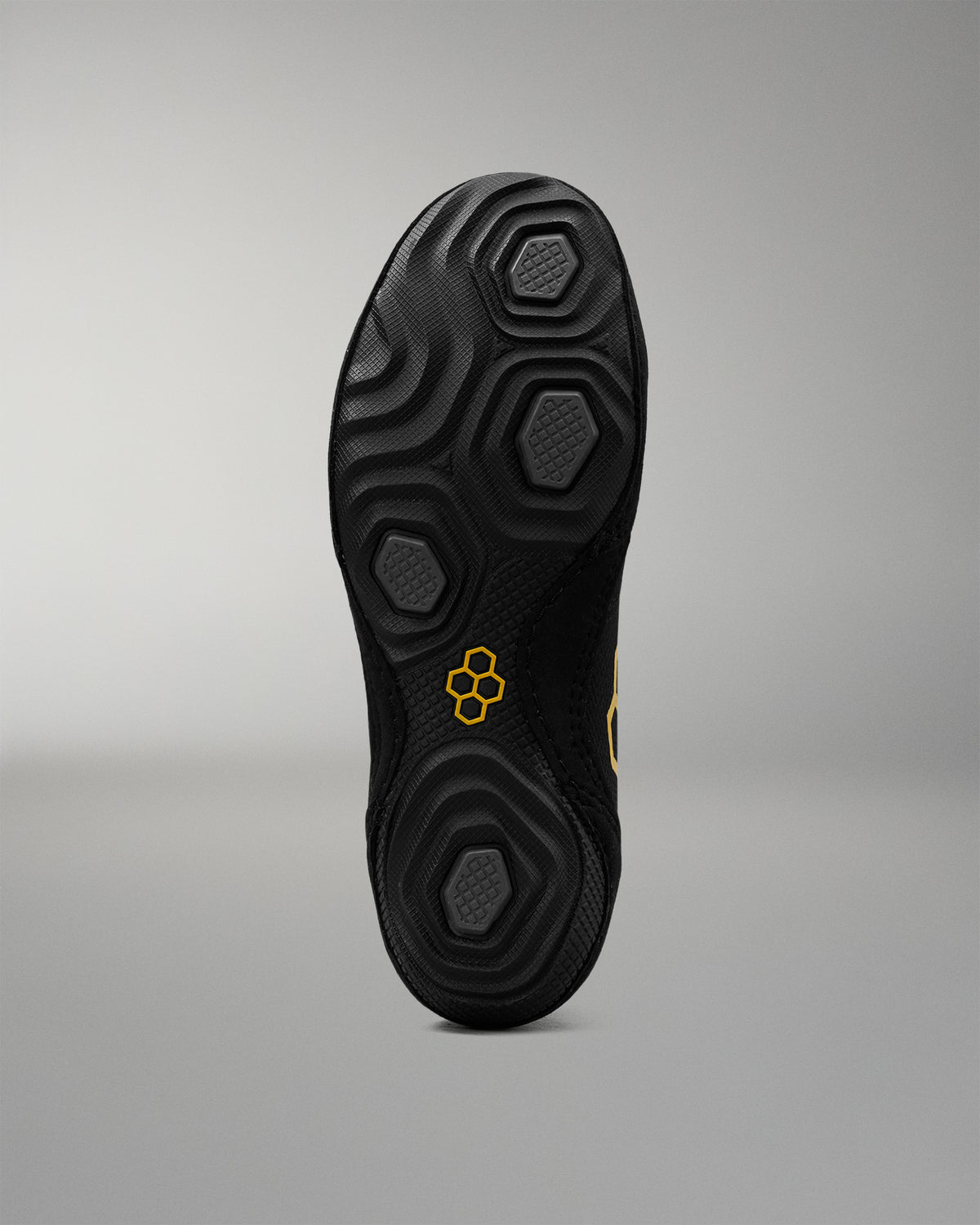 The black textured sole of a wrestling shoe, shown from a top-down view against a neutral gray background. The sole features a honeycomb-like tread pattern and a small yellow logo in the center.
