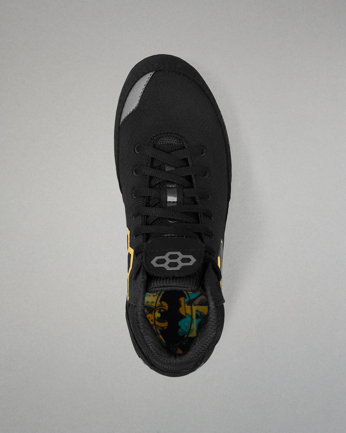 A black wrestling shoe shown from a top-down view on a neutral gray background. The shoe features a black mesh upper, black laces, a gray geometric logo on the tongue, and a colorful patterned lining visible at the collar.