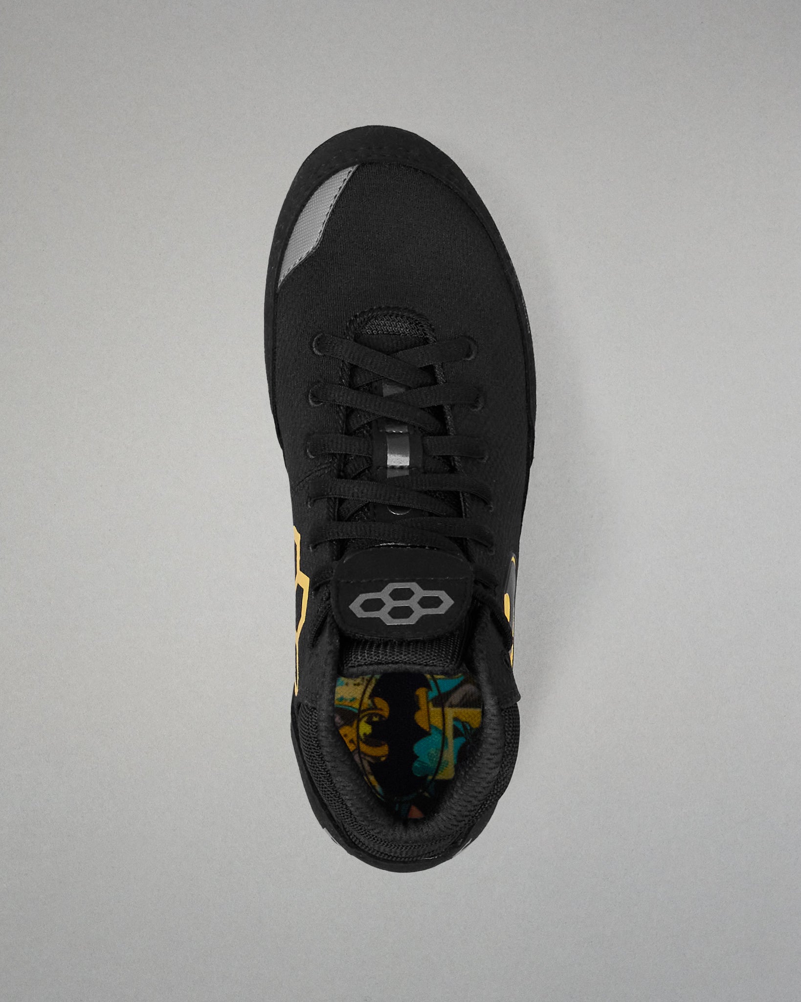 A black wrestling shoe shown from a top-down view on a neutral gray background. The shoe features a black mesh upper, black laces, a gray geometric logo on the tongue, and a colorful patterned lining visible at the collar.