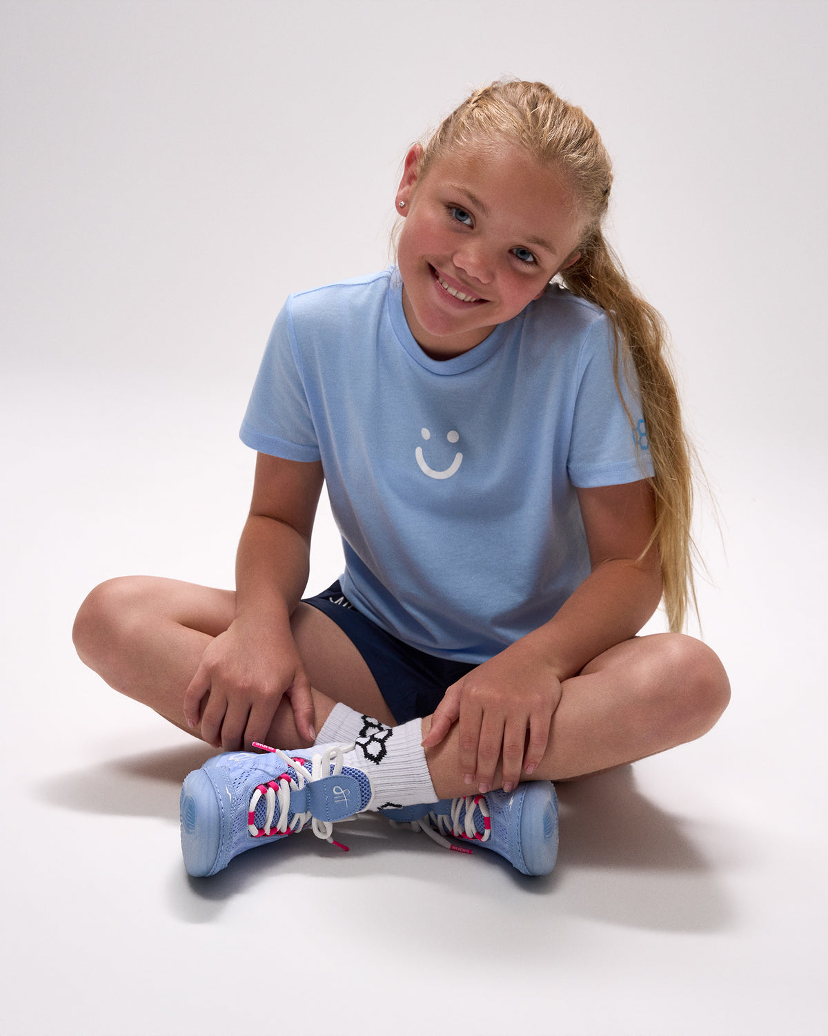 Young girl wearing a light blue t-shirt with a smiley face design, sitting on the floor with colorful socks.