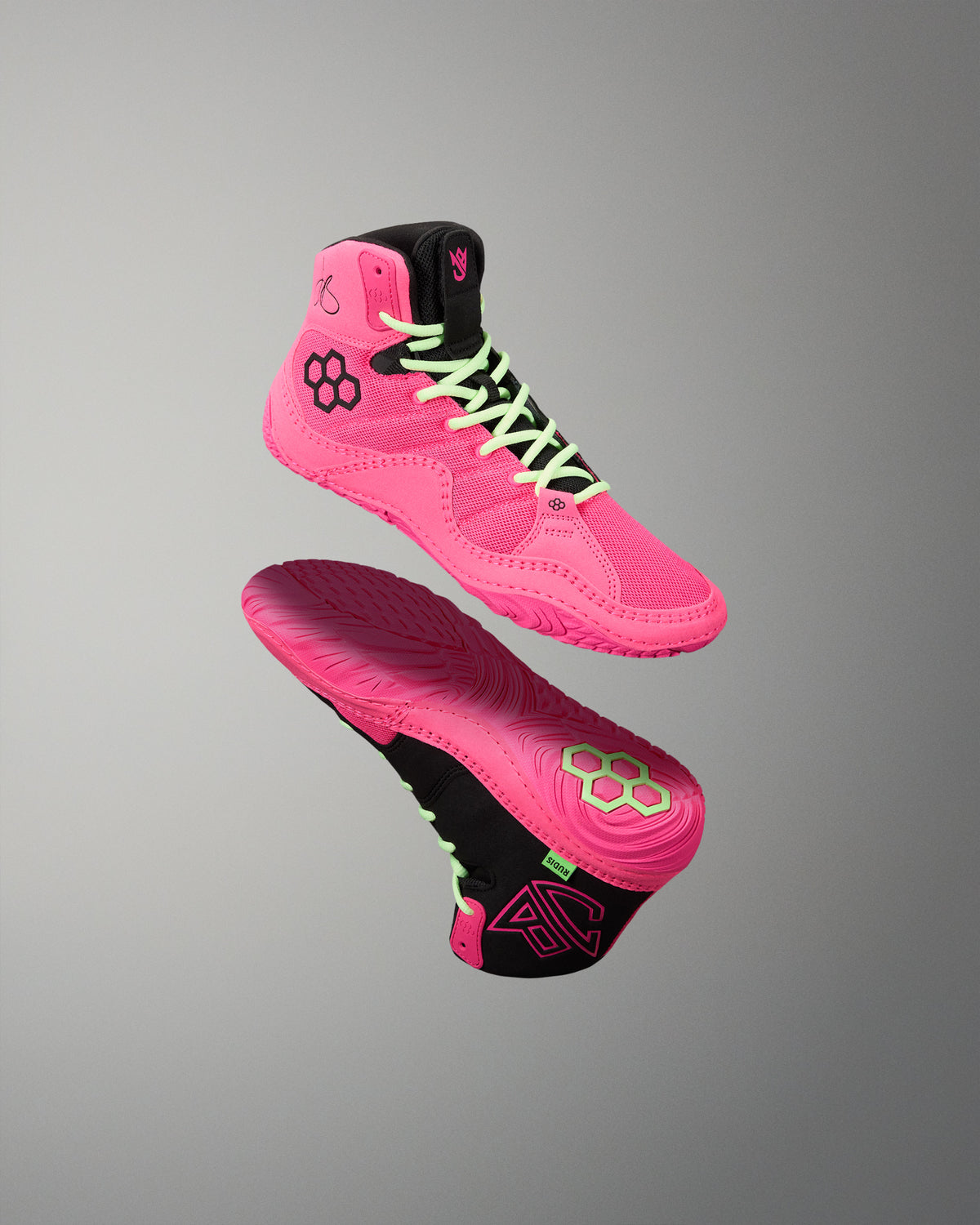 A pair of high-top athletic wrestling shoes in vibrant pink and black, shown floating against a neutral gray gradient background. One shoe is oriented to show the side profile with neon green laces, while the other shows the sole with a matching pink and black design.