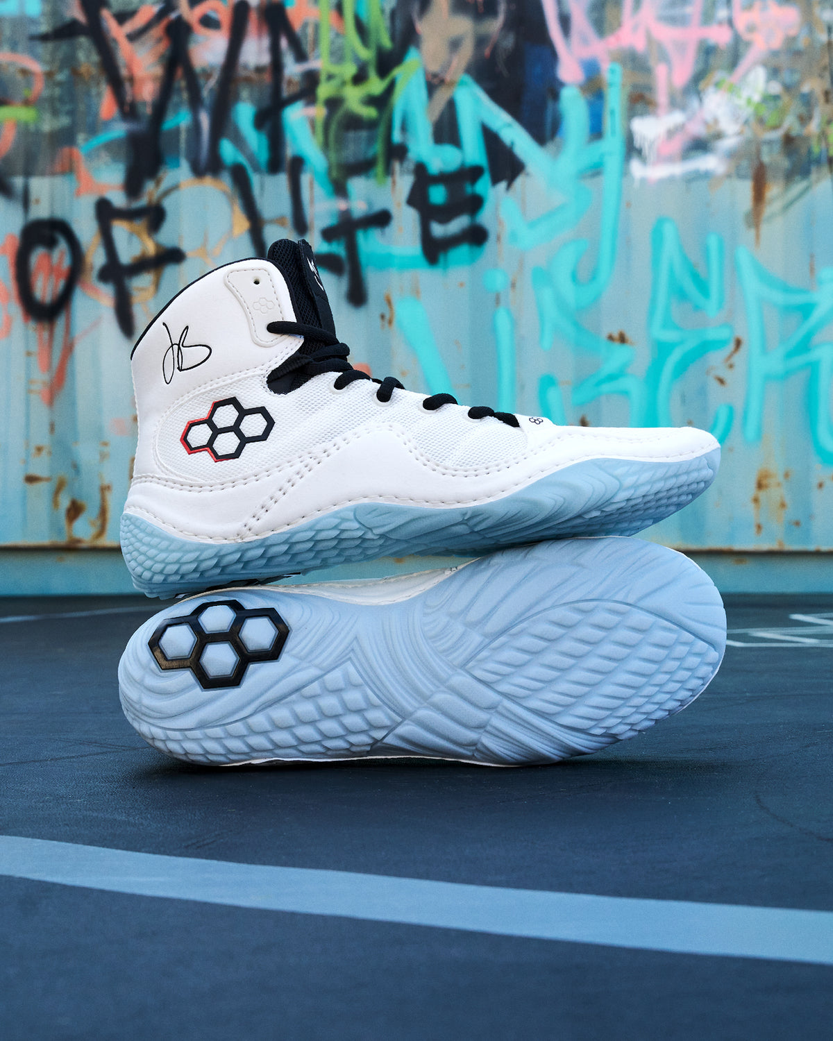 A pair of white high-top Wrestling shoes with black laces and a black-and-red geometric logo on the side, stacked with one shoe upright and the other showing the sole. The shoes are photographed outdoors against a graffiti-covered metal wall and dark pavement.