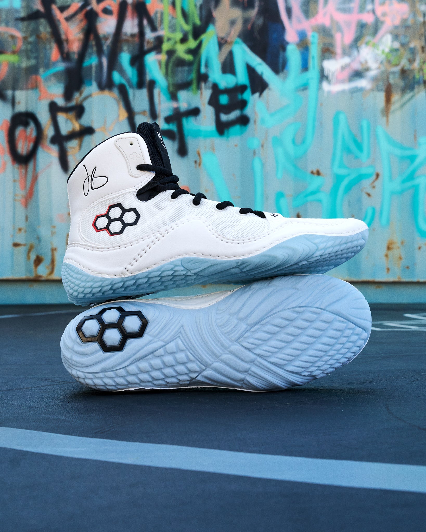 A pair of white high-top Wrestling shoes with black laces and a black-and-red geometric logo on the side, stacked with one shoe upright and the other showing the sole. The shoes are photographed outdoors against a graffiti-covered metal wall and dark pavement.
