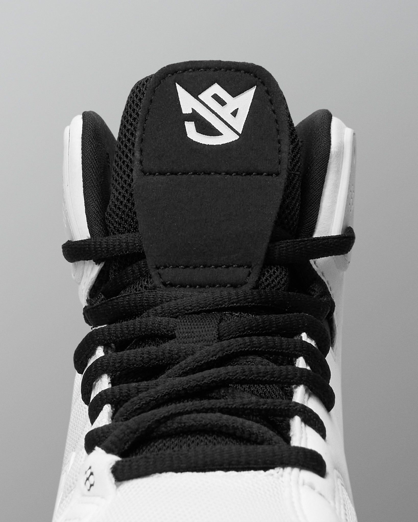 Close-up of a high-top wrestling shoe showing the black tongue patch with a white stylized logo, black laces, and white upper against a gray background.