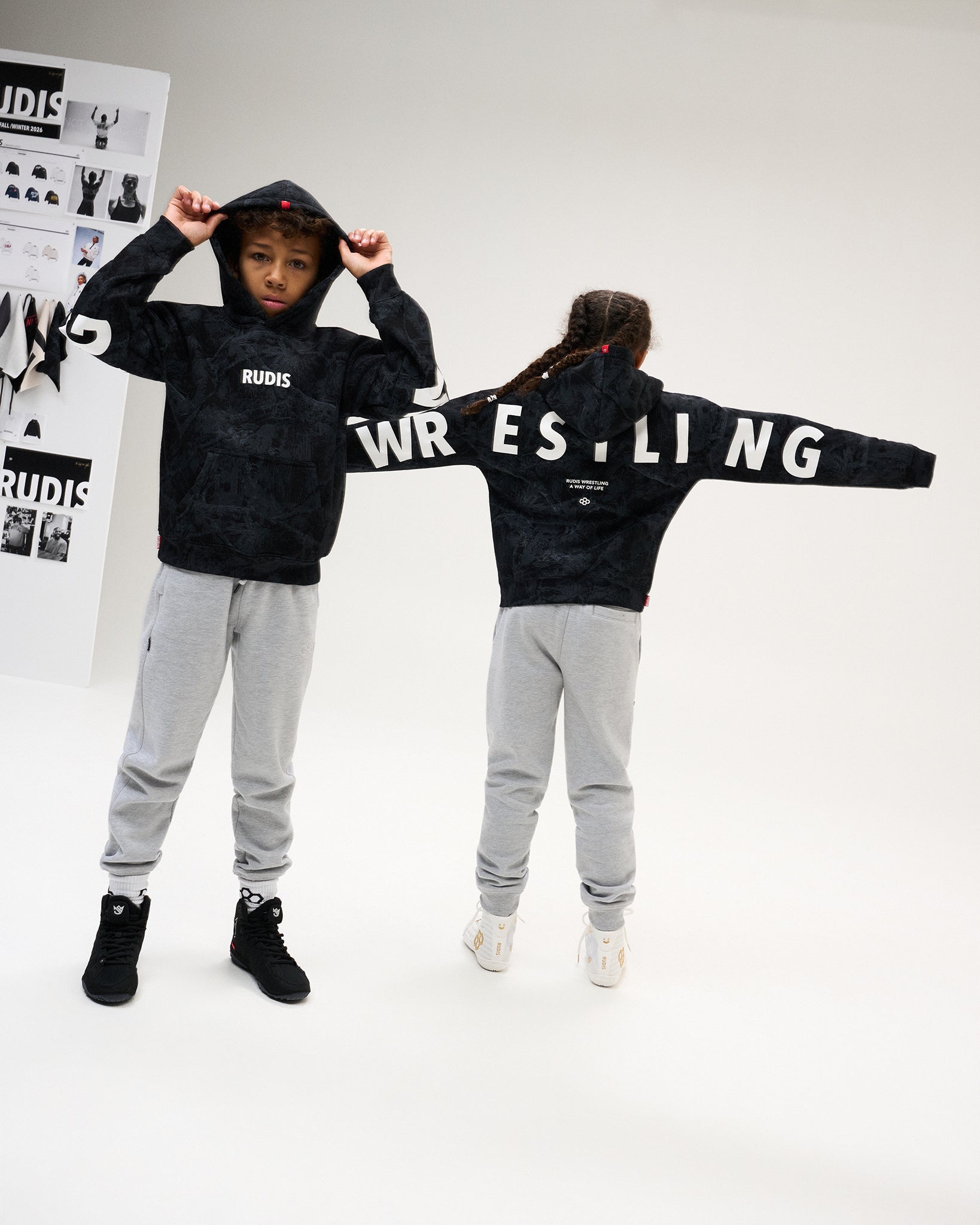 Two youth athletes model the RUDIS Wrestling Youth Relaxed Comfort Fleece Hoodie in Black Camo, showing the RUDIS chest logo and oversized WRESTLING graphics across the back and sleeves.