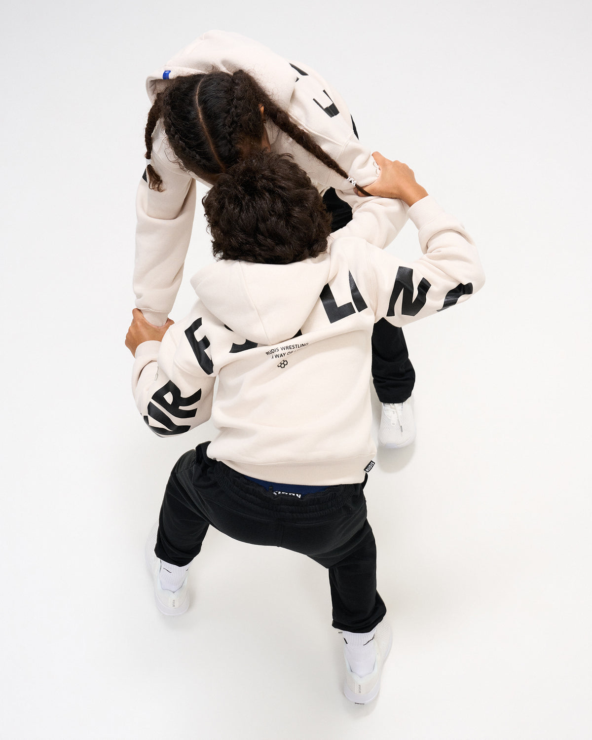 Overhead view of two youth athletes wearing RUDIS Wrestling Youth Relaxed Comfort Fleece Hoodies in Bone with contrasting black WRESTLING graphics across the back and sleeves.