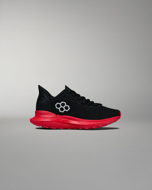 Side view of a black RUDIS training shoe featuring a red midsole and outsole with a white hexagon logo on the upper. Displayed on a neutral gray background.