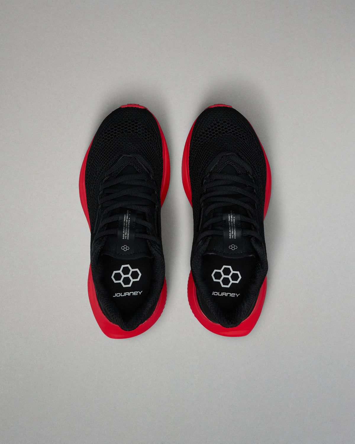 Top view of a pair of black RUDIS training shoes with red midsoles and black laces. The insoles display a white hexagon logo and the word ‘JOURNEY.’ Shoes are positioned side by side on a light gray background.
