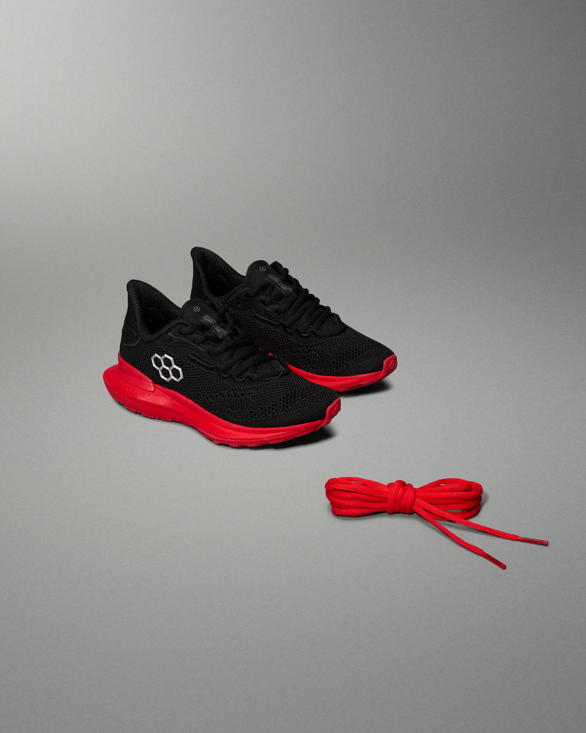 Angled view of a pair of black RUDIS training shoes with red midsoles and black laces, placed on a gray background. A separate pair of red replacement shoelaces is positioned in front of the shoes.