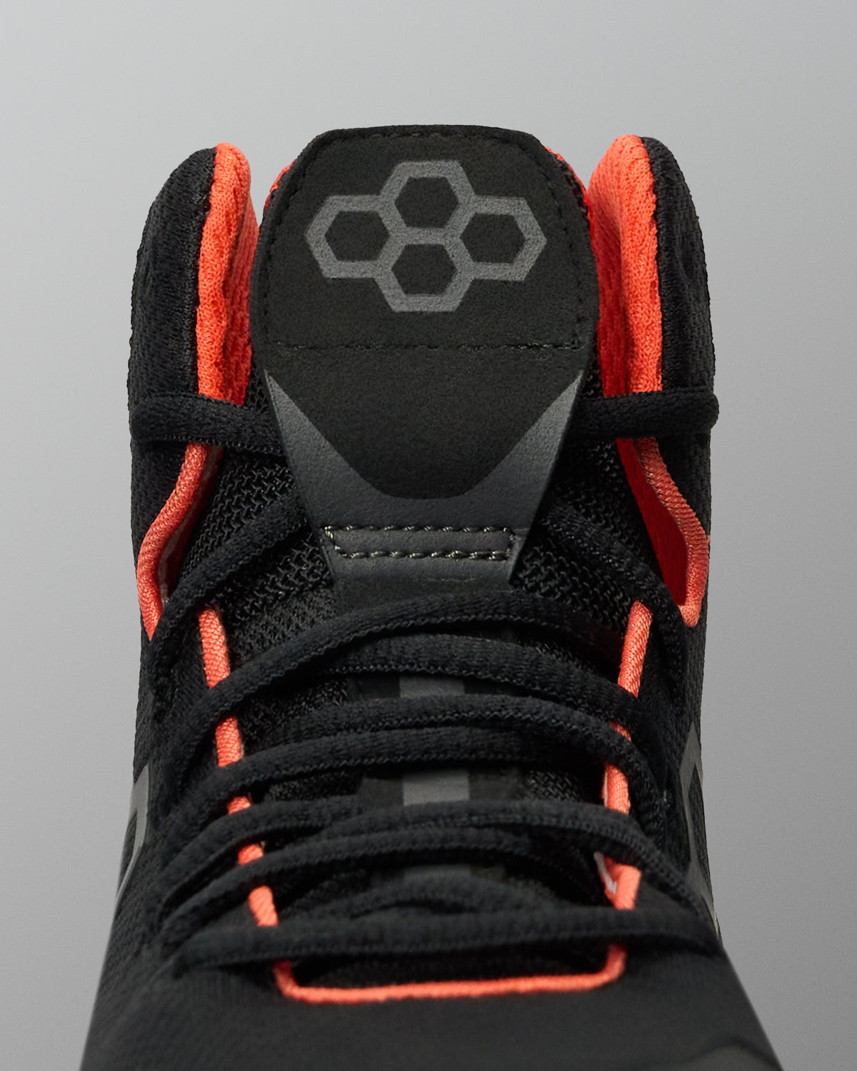 Close-up of a black high-top Wrestling Shoe with bright orange lining and laces, featuring a gray geometric logo on the tongue, set against a solid gray background.