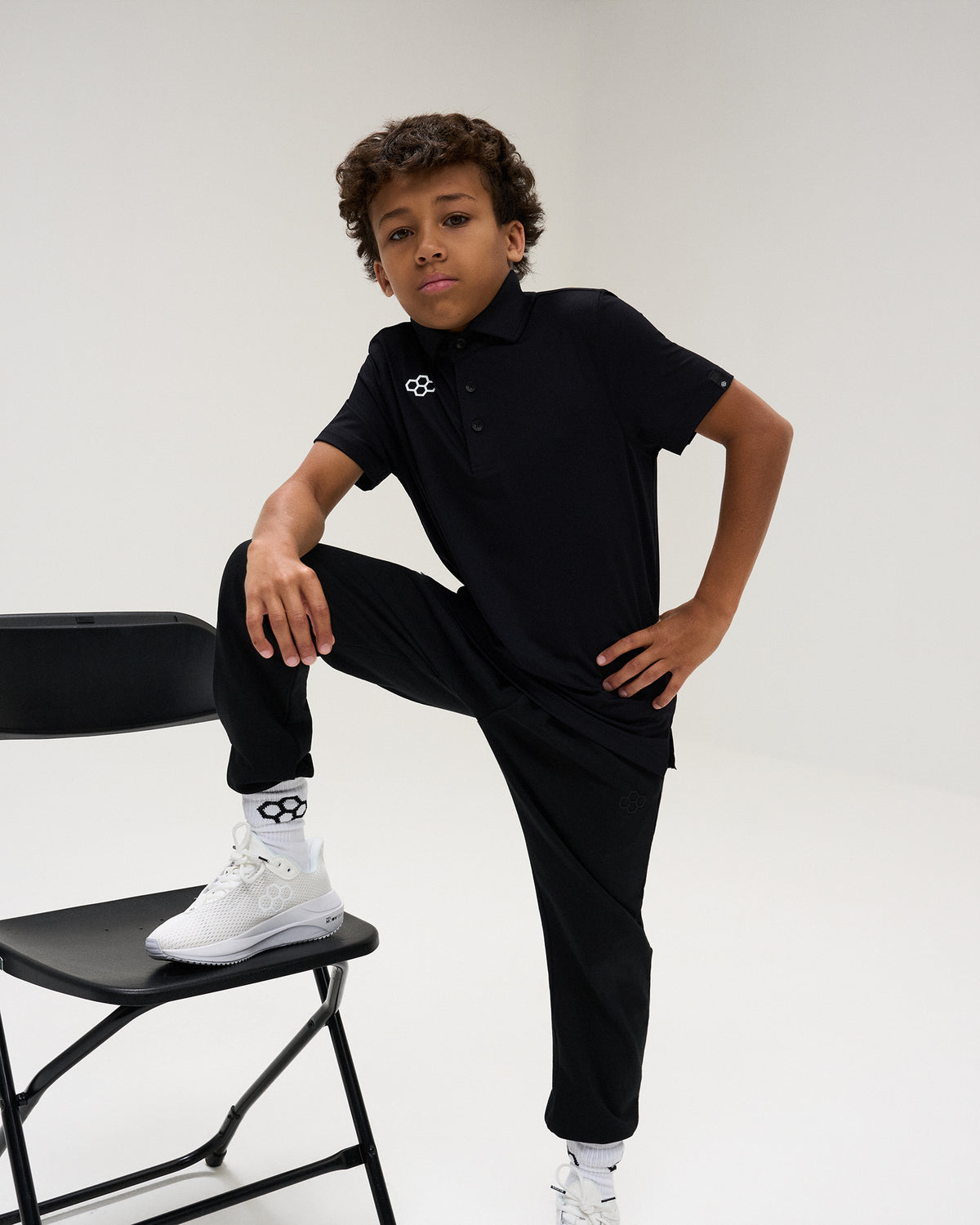 Youth athlete models the black RUDIS Textured Youth Classic Hex Polo and black athletic pants, posing with one foot on a chair.
