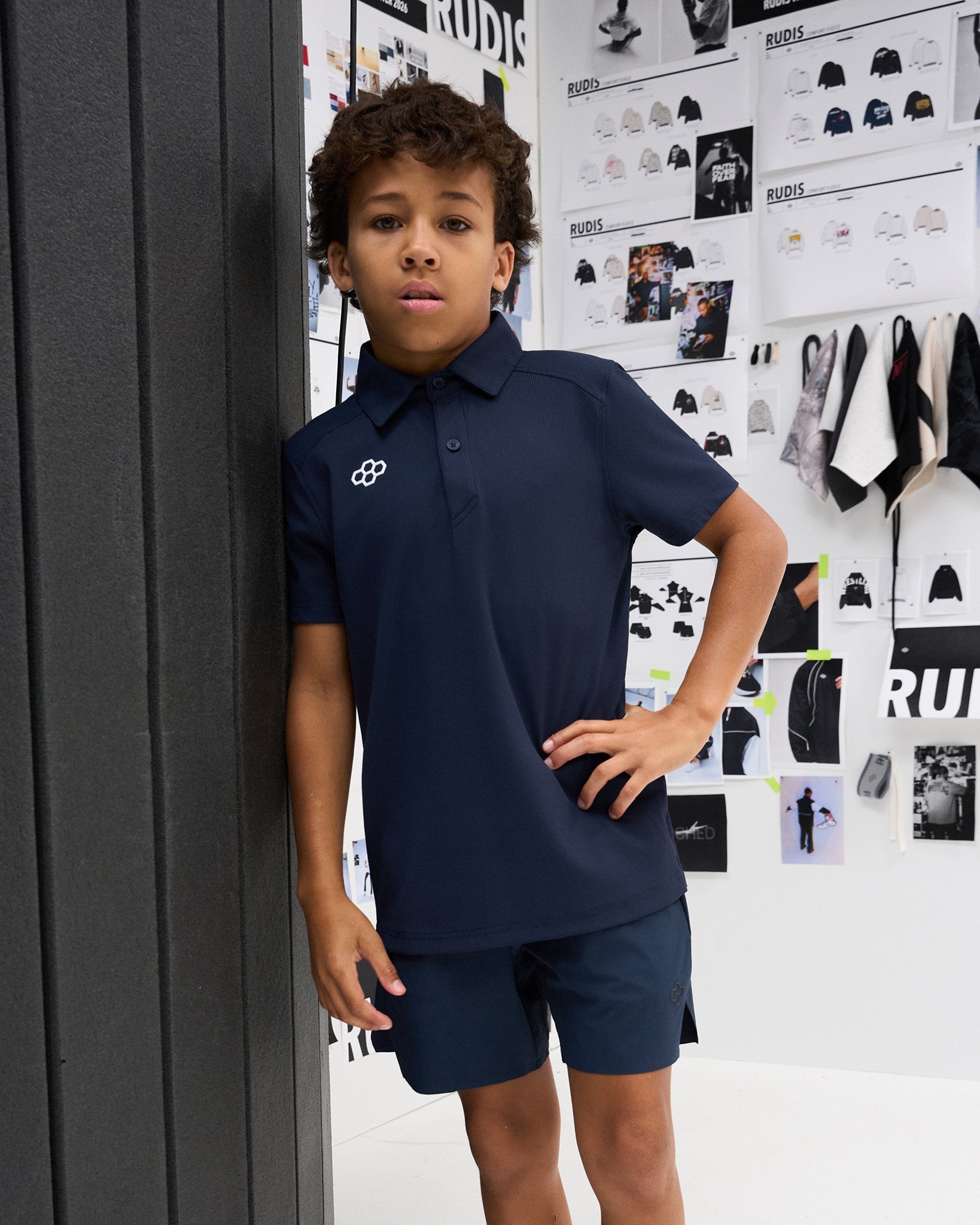 Youth athlete wears the navy RUDIS Textured Youth Classic Hex Polo with matching navy athletic shorts in a RUDIS design studio setting.