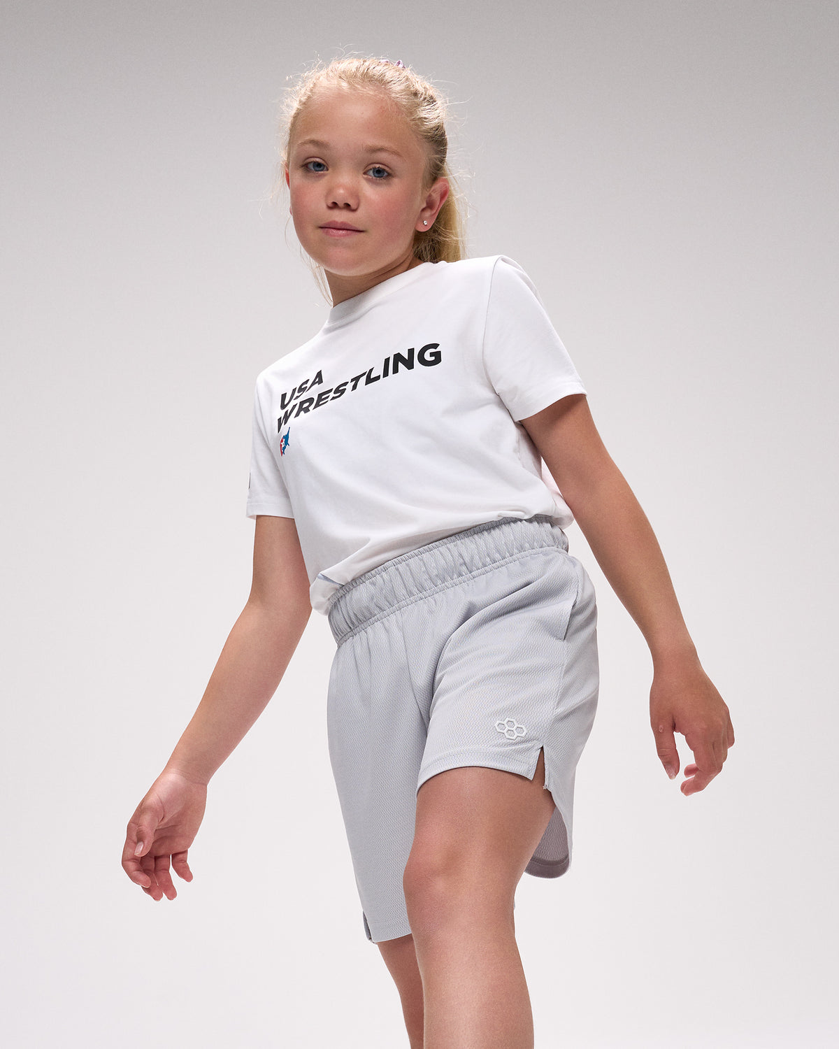 Child wearing a white 'USA Wrestling' t-shirt and gray shorts on a light gray background