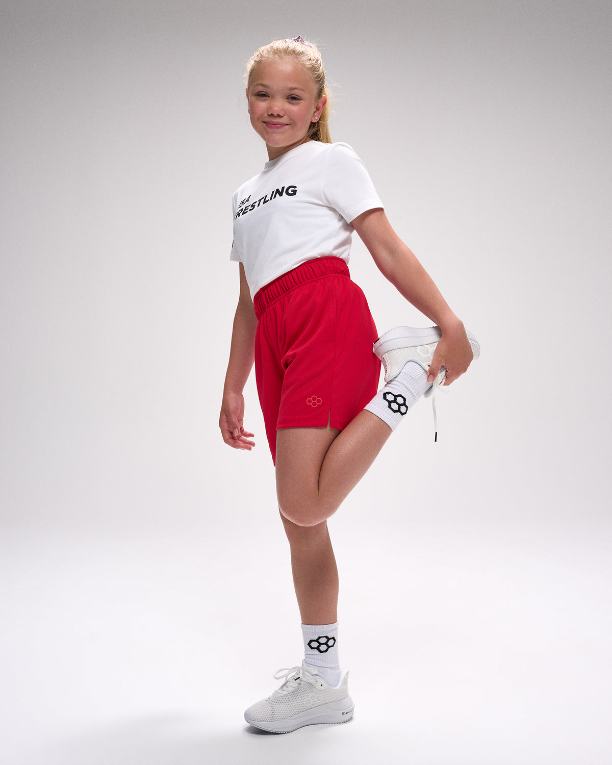 Young girl in white t-shirt with 'Wrestling' text, red shorts, and white sneakers on a gray background