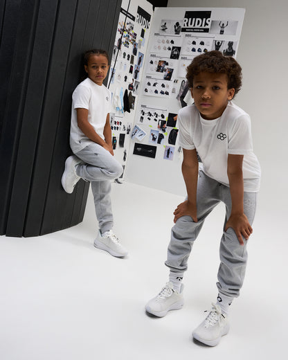 Two youth athletes model white RUDIS Essential Hex Youth T-Shirts with gray joggers and white athletic shoes in a RUDIS design studio.