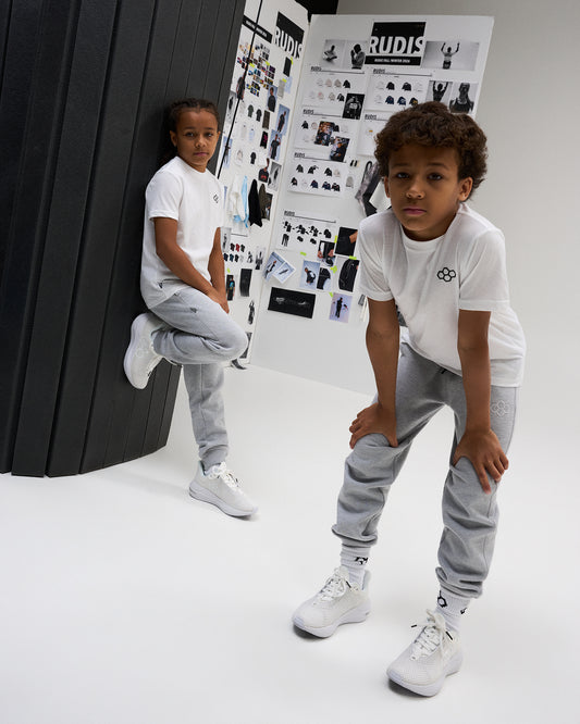 Two youth athletes model white RUDIS Essential Hex Youth T-Shirts with gray joggers and white athletic shoes in a RUDIS design studio.